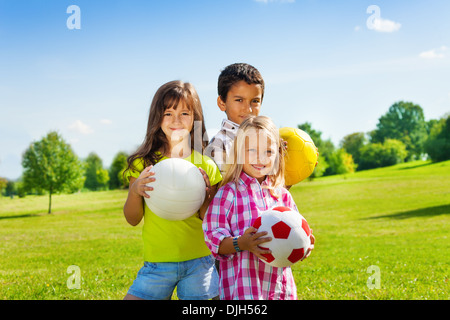 Team von drei glückliche Kinder, jungen und Mädchen stehen im sonnigen Sommerpark hält Sport Kugeln Stockfoto