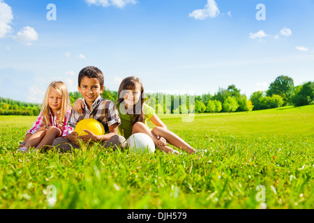 Drei glückliche Kinder, jungen und Mädchen sitzen im sonnigen Sommerpark hält Sport Kugeln Stockfoto