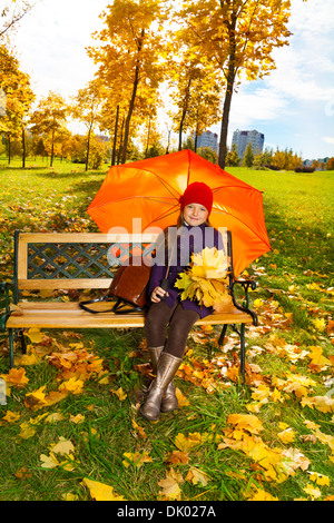 Glückliches kleines Mädchen hält Ahorn Blätter Blumenstrauß sitzt auf der Bank im Herbst Park unter Dach Stockfoto