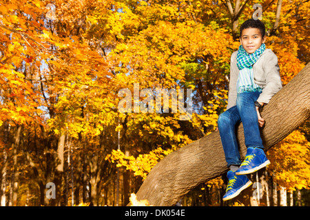 Schwarz 10 Jahre alten schwarzen Jungen sitzen auf dem Baum im Herbst Park mit Ahornblättern orange Stockfoto