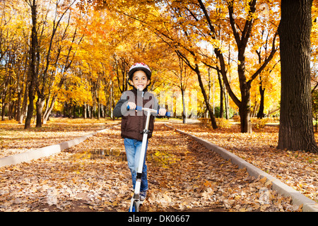 5 Jahre alten schwarzen Jungen tragen Helm fahren Roller im Herbst Park voller Laub Stockfoto