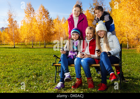 Fünf glückliche Schule Alter Kinder auf der Bank sitzen mit Mobiltelefonen glücklich und lächelnd, Jungen und Mädchen auf sonnigen Herbsttag warme Kleidung tragen Stockfoto