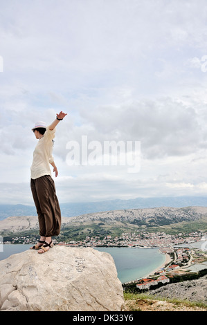 Mitte Erwachsene Frau mit ausgestreckten Armen auf Felsen mit Blick auf die Stadt Pag, Insel Pag, Kroatien, Europa Stockfoto