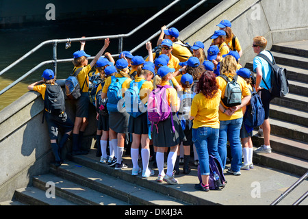 Kinder in blauen Mützen und farbenfrohen Schuluniformen für Jungen und Mädchen mit Lehrern, die an der Themse in Westminster London, England, teilhaben Stockfoto