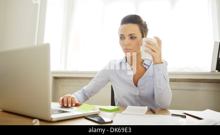 Hübsche junge Geschäftsfrau hält eine Tasse Kaffee auf Laptop-Computer arbeiten. Schöne junge Frau sitzt am Schreibtisch arbeiten Stockfoto