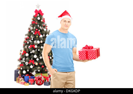 Hübscher Kerl mit Weihnachtsmütze hält ein Geschenk und posiert vor einem Weihnachtsbaum Stockfoto