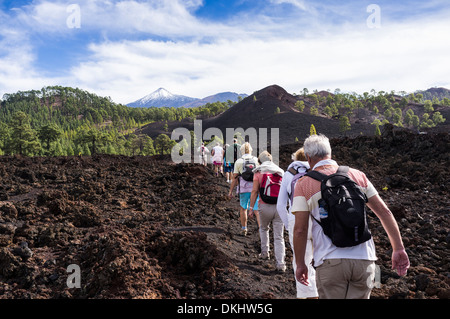 Eine Gruppe von Wanderern im Gänsemarsch Kreuz einen felsigen Lavastrom im Chinyero mit einer schneebedeckten Teide im Hintergrund, Teneriffa. Stockfoto