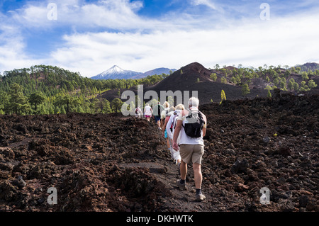 Eine Gruppe von Wanderern im Gänsemarsch Kreuz einen felsigen Lavastrom im Chinyero mit einer schneebedeckten Teide im Hintergrund, Teneriffa. Stockfoto