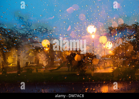 Blick durch die Regentropfen auf ein Fenster zur Straße draußen in der Morgendämmerung mit Straßenlaternen Abfackeln durch den Regen Stockfoto