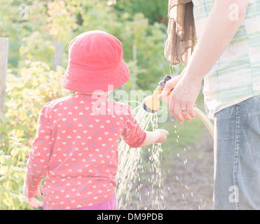 Lifestyle Sommer-Szene. Kleine Mädchen spielen mit Eltern im Garten, Gefühl Wasser aus Sprinkleranlagen Stockfoto
