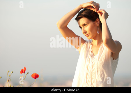 Junge Frau im Kleid steht bei Sonnenuntergang in einem Maisfeld mit Mohnblumen Stockfoto