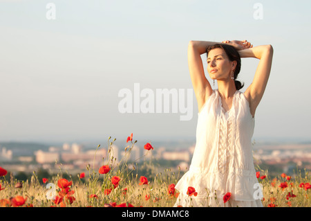 Junge Frau im Kleid steht bei Sonnenuntergang in einem Maisfeld mit Mohnblumen Stockfoto