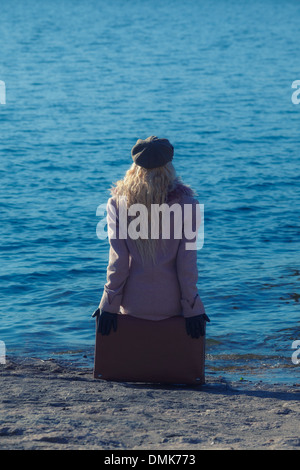 eine Frau in einem rosa Mantel sitzt auf einem Koffer am Meer Stockfoto