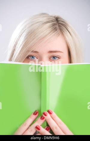 Hübsche blonde Teenager peeking über ein grünes Buch hielt vor ihr Gesicht. Stockfoto