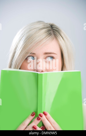 Hübsche blonde Teenager peeking über ein grünes Buch hielt vor ihr Gesicht. Stockfoto