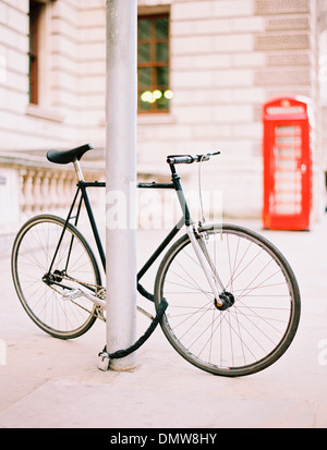 Ein Fahrrad gefesselt und an einen Laternenpfahl auf einer Londoner Straße gesperrt. Eine rote öffentliche Telefonzelle im Hintergrund. Stockfoto