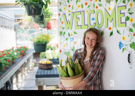 Im Sommer auf einem Bio-Bauernhof. Ein Mädchen hält einen Korb mit frischem Mais durch Willkommensschild. Stockfoto