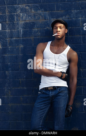 Schöner schwarzer Mann, Afroamerikaner, Rauchen, trägt blaue Jeans und Zeitungsjunge Mütze Wand gelehnt, eine Vintage Mode-Konzept Stockfoto