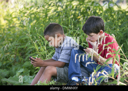 Zwei Jungs sitzen in einem Feld eine auf ein Smartphone und ein mit einem digital-Tablette. Stockfoto