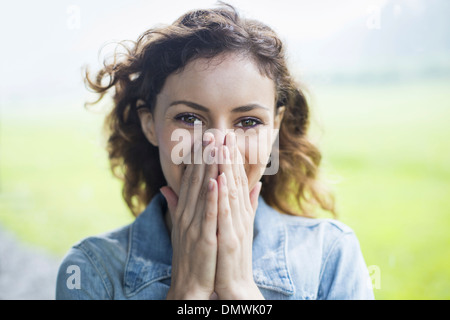 Eine junge Frau in einer ländlichen Landschaft mit vom Wind verwehten lockiges Haar. Bedeckte ihr Gesicht mit den Händen und lacht. Stockfoto
