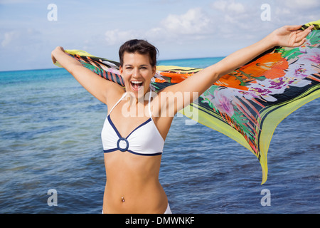 Temperamentvolle Frau Lachen im Bikini am Meer mit einem bunt gemusterten Schal in ihren ausgestreckten Händen flattern im Wind gegen einen Ozean Hintergrund Stockfoto