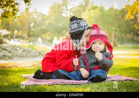 Kleines Mädchen flüstert ein Geheimnis um ihr Baby Bruder tragen Wintermäntel und Hüte sitzen draußen im Park. Stockfoto