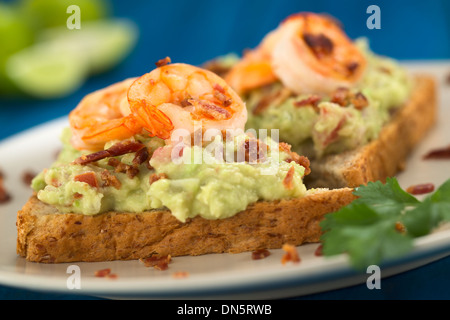 Vollkorn Toast Brotscheiben mit Guacamole, gebratene Garnelen und gebratenen Speckwürfeln serviert auf Platte auf blau Holz Stockfoto