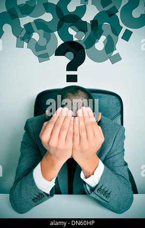 ein Geschäftsmann in einem Schreibtisch mit seinen Händen in seinem Kopf und eine Wolke von Fragezeichen über ihm sitzen Stockfoto