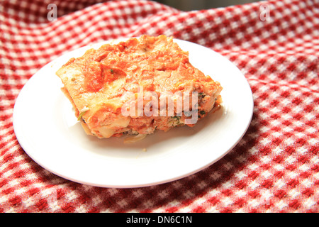 Ein Stück hausgemachte Fleisch Lasagne auf einem weißen Teller mit einer Flasche Rotwein im Hintergrund auf einer karierten Tischdecke Stockfoto