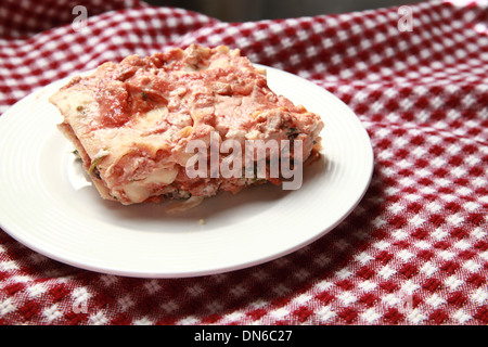 Ein Stück hausgemachte Fleisch Lasagne auf einem weißen Teller mit einer Flasche Rotwein im Hintergrund auf einer karierten Tischdecke Stockfoto