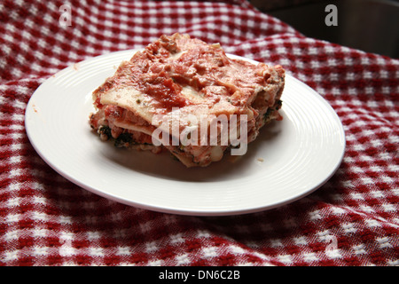 Ein Stück hausgemachte Fleisch Lasagne auf einem weißen Teller mit einer Flasche Rotwein im Hintergrund auf einer karierten Tischdecke Stockfoto