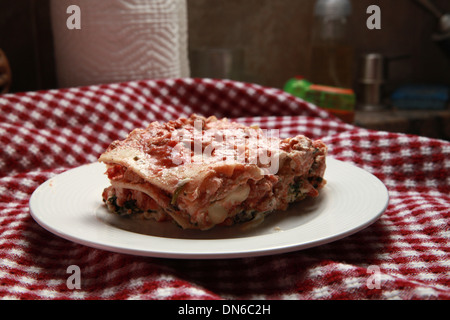 Ein Stück hausgemachte Fleisch Lasagne auf einem weißen Teller mit einer Flasche Rotwein im Hintergrund auf einer karierten Tischdecke Stockfoto
