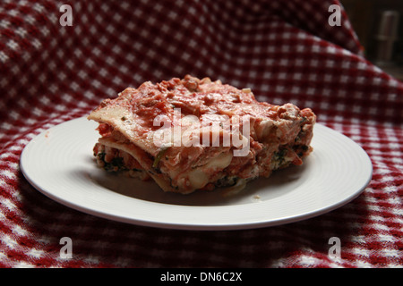 Ein Stück hausgemachte Fleisch Lasagne auf einem weißen Teller mit einer Flasche Rotwein im Hintergrund auf einer karierten Tischdecke Stockfoto