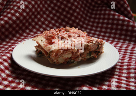 Ein Stück hausgemachte Fleisch Lasagne auf einem weißen Teller mit einer Flasche Rotwein im Hintergrund auf einer karierten Tischdecke Stockfoto