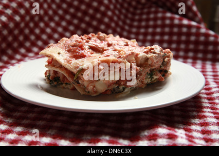 Ein Stück hausgemachte Fleisch Lasagne auf einem weißen Teller mit einer Flasche Rotwein im Hintergrund auf einer karierten Tischdecke Stockfoto