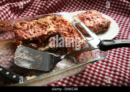 Ein Stück hausgemachte Fleisch Lasagne auf einem weißen Teller mit einer Flasche Rotwein im Hintergrund auf einer karierten Tischdecke Stockfoto