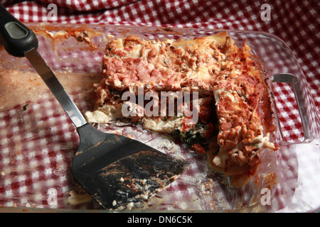 Ein Stück hausgemachte Fleisch Lasagne auf einem weißen Teller mit einer Flasche Rotwein im Hintergrund auf einer karierten Tischdecke Stockfoto