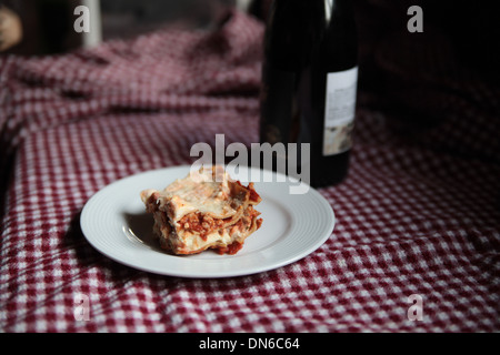 Ein Stück hausgemachte Fleisch Lasagne auf einem weißen Teller mit einer Flasche Rotwein im Hintergrund auf einer karierten Tischdecke Stockfoto