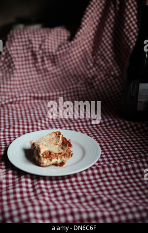 Ein Stück hausgemachte Fleisch Lasagne auf einem weißen Teller mit einer Flasche Rotwein im Hintergrund auf einer karierten Tischdecke Stockfoto