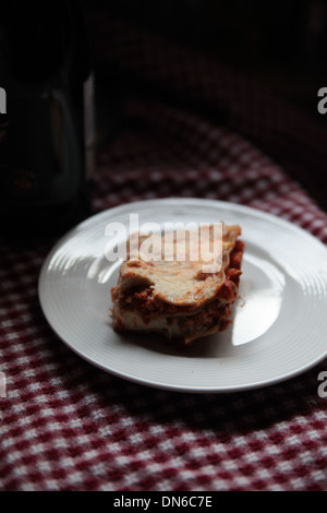 Ein Stück hausgemachte Fleisch Lasagne auf einem weißen Teller mit einer Flasche Rotwein im Hintergrund auf einer karierten Tischdecke Stockfoto