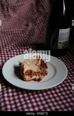 Ein Stück hausgemachte Fleisch Lasagne auf einem weißen Teller mit einer Flasche Rotwein im Hintergrund auf einer karierten Tischdecke Stockfoto