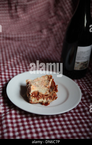 Ein Stück hausgemachte Fleisch Lasagne auf einem weißen Teller mit einer Flasche Rotwein im Hintergrund auf einer karierten Tischdecke Stockfoto