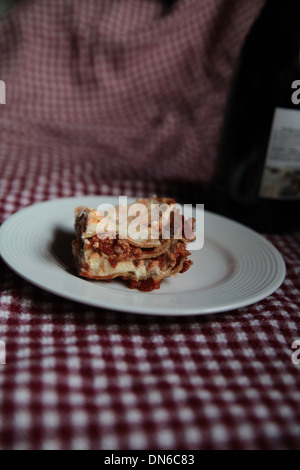 Ein Stück hausgemachte Fleisch Lasagne auf einem weißen Teller mit einer Flasche Rotwein im Hintergrund auf einer karierten Tischdecke Stockfoto