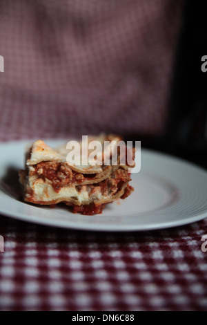 Ein Stück hausgemachte Fleisch Lasagne auf einem weißen Teller mit einer Flasche Rotwein im Hintergrund auf einer karierten Tischdecke Stockfoto