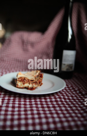 Ein Stück hausgemachte Fleisch Lasagne auf einem weißen Teller mit einer Flasche Rotwein im Hintergrund auf einer karierten Tischdecke Stockfoto