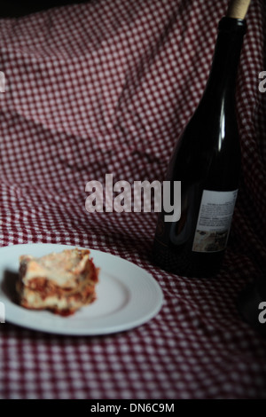Ein Stück hausgemachte Fleisch Lasagne auf einem weißen Teller mit einer Flasche Rotwein im Hintergrund auf einer karierten Tischdecke Stockfoto