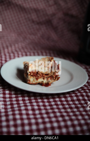 Ein Stück hausgemachte Fleisch Lasagne auf einem weißen Teller mit einer Flasche Rotwein im Hintergrund auf einer karierten Tischdecke Stockfoto