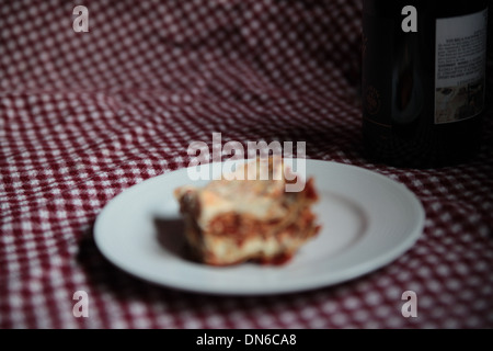 Ein Stück hausgemachte Fleisch Lasagne auf einem weißen Teller mit einer Flasche Rotwein im Hintergrund auf einer karierten Tischdecke Stockfoto