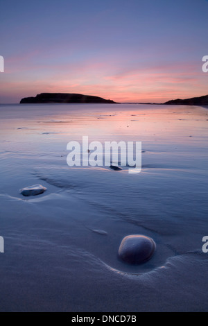 Kieselsteine und nassen Sand links von der zurückweichenden Flut am Strand von Rhossili Bay bei Sonnenuntergang. Burry Holms können in der Ferne gesehen werden. Stockfoto