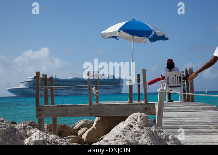 Das Kreuzfahrtschiff liegt vor der Bahaman-Insel Eleuthera vor Anker. Ein Rettungsschwimmer sitzt unter einem blau-weißen Schirm auf einem kleinen Pier am Ufer. Stockfoto